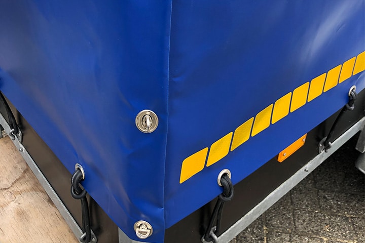 Detailfoto van hoeksluiting en gele veiligheidsmarkeringstape op een aanhangerafdekzeil. Hoewel niet verplicht, draagt de toevoeging van de markeringstape bij aan een verhoogde zichtbaarheid en veiligheid tijdens het gebruik van de aanhanger.